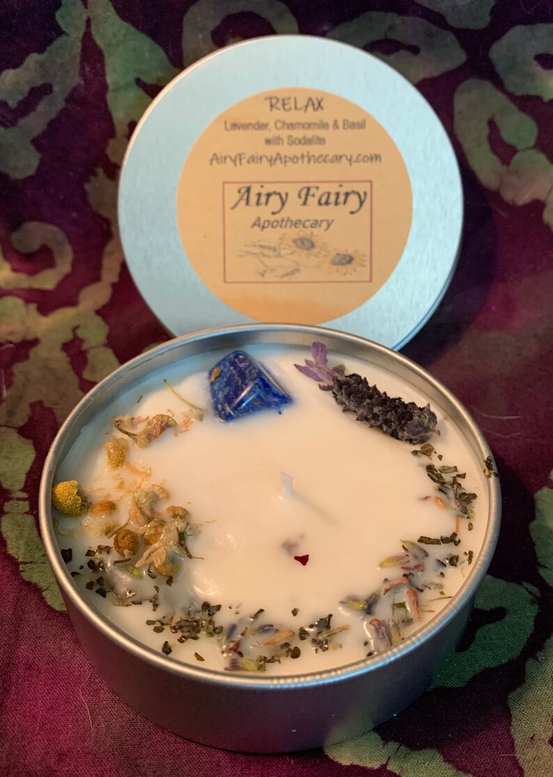 May include: Airy Fairy Apothecary candle in a silver tin with a white wax base. The candle has a blue sodalite crystal, dried lavender, and chamomile flowers embedded in the wax. The label on the tin reads "Relax Lavender, Chamomile & Basil with Sodalite AiryFairyApothecary.com Airy Fairy Apothecary".