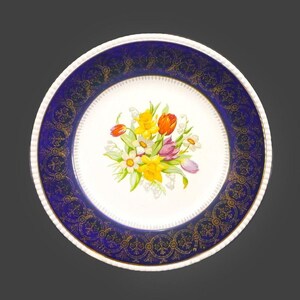 May include: A white ceramic plate with a blue and gold rim. The centre of the plate features a bouquet of yellow daffodils, pink and purple tulips, and white lily of the valley flowers.