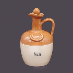 Könnte beinhalten: Eine Keramikkanne mit brauner Glasur und weißem Boden. Die Kanne hat einen Henkel und einen Deckel mit einem Knopf oben. Die Kanne ist mit "Rum" in schwarzen Buchstaben beschriftet.