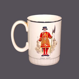 Könnte beinhalten: Weiße Keramiktasse mit silbernem Rand. Die Tasse zeigt eine farbenfrohe Illustration eines Beefeater-Wächters in roter Uniform, der eine Lanze hält. Der Text "RIGRAW HAMOY" ist auf der Unterseite der Tasse aufgedruckt.