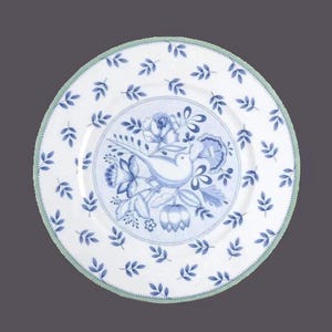 May include: A white ceramic dinner plate with a blue and green floral design. The centre of the plate features a stylised bird surrounded by flowers and leaves. The rim of the plate is decorated with a repeating pattern of blue leaves.