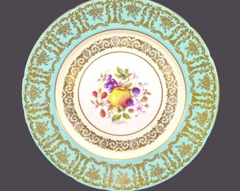 May include: A white porcelain plate with a gold rim and a turquoise border. The center of the plate features a hand-painted design of fruit, including apples, grapes, and berries.