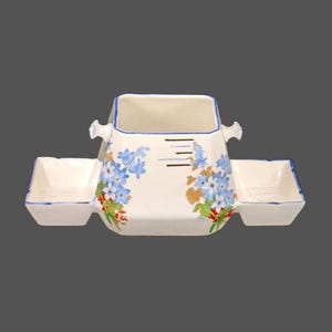 Könnte beinhalten: Eine weiße Keramik-Servierplatte mit einem quadratischen Mittelteil und zwei kleineren rechteckigen Seitenteilen. Der Mittelteil hat ein Blumenmuster mit blauen Blüten und grünen Blättern. Die Seitenteile haben einen blauen Rand.