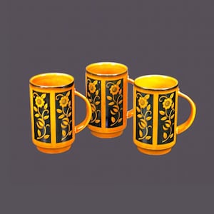 Op de afbeelding: Drie oranje keramische mokken met zwarte bloemenmotieven. De mokken hebben een gouden rand en staan op een grijs oppervlak.