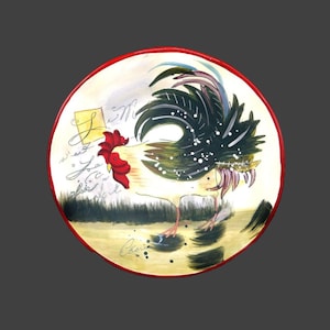 May include: A round ceramic plate with a red rim features a hand-painted design of a rooster with a black, green, and white tail. The rooster is standing on a green patch of grass with black and white splatters of paint. The plate has French text written on it.