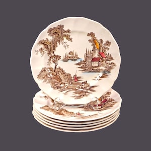 May include: Set of six white ceramic dinner plates with a brown and red landscape design featuring a cottage and trees. The plates have a scalloped edge.