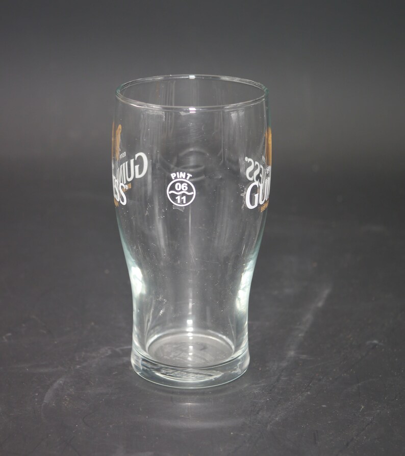 May include: A clear glass pint glass with the Guinness logo printed on the side. The logo is in gold and black and includes the text "Guinness" and "Dublin, Ireland". The glass also has a white circle with the text "Pint 0.6 11" printed on the side.