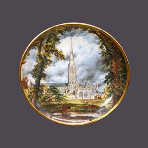 May include: A round ceramic plate with a gold rim features a painted scene of a cathedral with a tall spire, surrounded by trees and a grassy field with cows. The sky is painted with clouds.