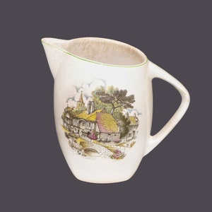 May include: A white ceramic pitcher with a green rim and a brown, green, and yellow illustration of a cottage with a thatched roof and a bridge. The pitcher has a handle.