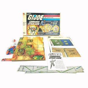 Può includere: Un gioco da tavolo vintage G.I. Joe Commando Attack Game nella sua scatola originale. Il gioco include un tabellone di gioco, pezzi da gioco, carte e istruzioni. La scatola presenta un'illustrazione colorata di personaggi G.I. Joe in azione.