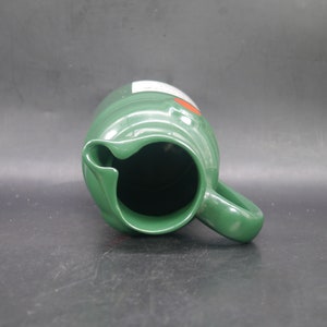 May include: A green ceramic pitcher with a handle. The pitcher has a white label with red lettering.