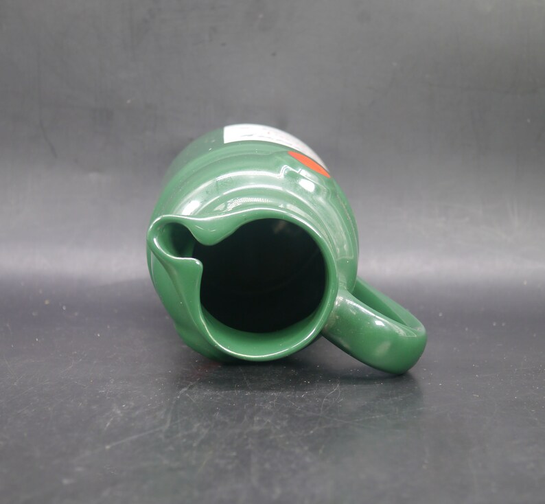 May include: A green ceramic pitcher with a handle. The pitcher has a white label with red lettering.