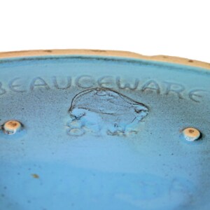 以下が含まれることがあります： 薄い青色のセラミックボウルで、底に「BEAUCEWARE」という文字が刻まれています。ボウルは薄い茶色の縁取りがあり、底に2つの小さな丸い薄い茶色の点があります。