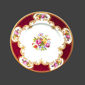 May include: A white porcelain dinner plate with a red and gold rim. The plate features a floral design with pink, yellow, and blue flowers.