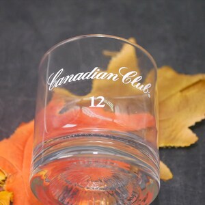 May include: A clear glass tumbler with the words "Canadian Club" and the number "12" etched on the side. The tumbler is sitting on a bed of orange and yellow autumn leaves.