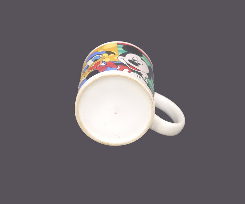 May include: A white ceramic mug with a colorful cartoon design featuring Mickey Mouse, Donald Duck, and Goofy. The mug has a handle and a white interior.
