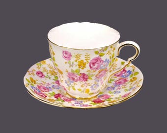 Pode incluir: Uma chávena de chá e um pires brancos com um padrão floral em tons de rosa, amarelo e azul. A chávena tem uma borda dourada e o pires tem uma borda ondulada.