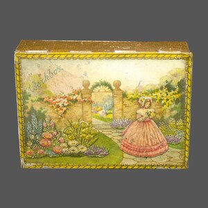 Peut inclure: Une boîte à ouvrage vintage avec une bordure dorée et une image colorée d'une femme dans un jardin. L'image représente un mur de briques, une porte, des fleurs et un chemin. Le texte "Workbox" est imprimé sur la boîte.