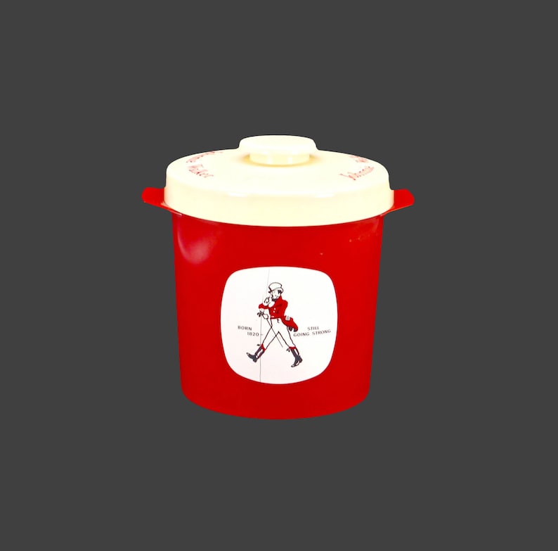 May include: Red plastic container with a white lid and a Johnnie Walker logo featuring a man in a red coat walking with a stick. The logo says "Born 1820 Still Going Strong".