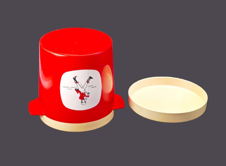 May include: A red plastic container with a white label that says "DOUBLE YOUR ENJOYMENT" and a picture of a person upside down. The container has a white lid.