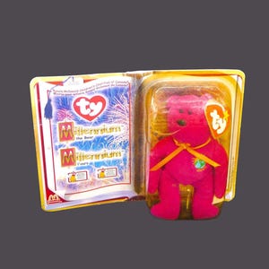 May include: A pink Ty Beanie Baby called Millennium the Bear, wearing a yellow ribbon with a globe design. The bear is packaged in a clear plastic case with a gold and white cardboard backing. The backing features the Ty logo and the words "Millennium the Bear" and "Ronald McDonald's Children's Charities of Canada."
