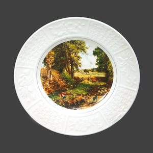 May include: White ceramic plate with a raised rim and a detailed embossed design. The center of the plate features a colorful landscape painting of a wooded area with a path leading through it.