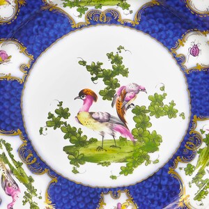 Puede incluir: Un plato de porcelana blanca con un borde azul y dorado. El centro del plato presenta un dise&ntilde;o pintado a mano de dos p&aacute;jaros posados en una rama con hojas verdes. Los p&aacute;jaros son rosas, amarillos y marrones.