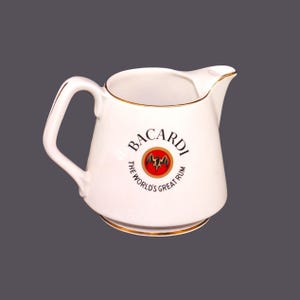 Può includere: Brocca in ceramica bianca con bordo dorato e logo Bacardi. Il logo presenta un pipistrello e il testo "Bacardi, The World's Great Rum".