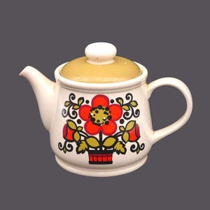 May include: A white ceramic teapot with a floral design. The teapot has a yellow lid and a red flower with green leaves and a black centre. The flower is in a red and black pot.