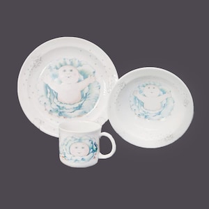 May include: A white ceramic children's dinnerware set featuring a Cabbage Patch Kid design. The set includes a plate, bowl, and mug.