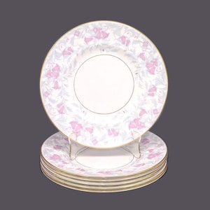 May include: A set of six white porcelain dinner plates featuring a delicate floral design in shades of pink and grey. The plates have a gold rim and are stacked on top of each other.