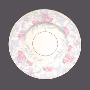 Op de afbeelding: Een wit keramiek bord met een gouden rand en een bloemenmotief. Het ontwerp heeft roze bloemen en blauwe bladeren op een witte achtergrond.