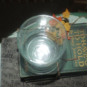 May include: A clear glass cup sits on top of a stack of vintage books. The books have a green cover with gold lettering that reads "To Have & To Hold" by Mary Johnston. The top book has a brown paper label with the word "TED" printed on it.