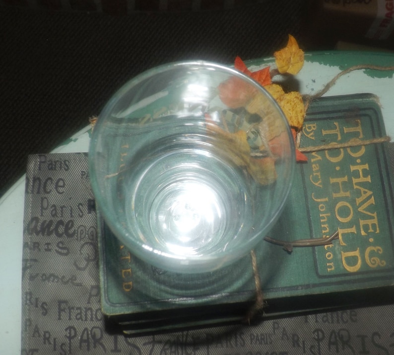 May include: A clear glass cup sits on top of a stack of vintage books. The books have a green cover with gold lettering that reads "To Have & To Hold" by Mary Johnston. The top book has a brown paper label with the word "TED" printed on it.