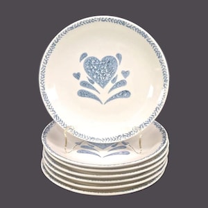 May include: A stack of eight white ceramic dinner plates with a blue heart design. The plates have a blue and white border.