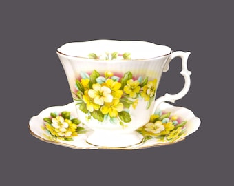 May include: A white teacup and saucer with a floral pattern featuring yellow primrose flowers and green leaves. The cup has a scalloped rim and a delicate handle. The saucer has a matching floral design.