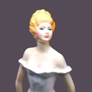 May include: A porcelain figurine of a woman with blonde hair, wearing a white dress with blue floral accents. The figurine has a delicate and elegant design.