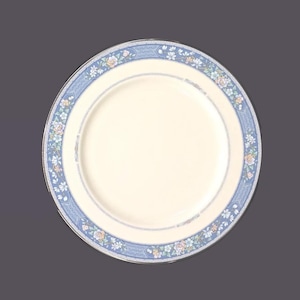 Può includere: Un piatto rotondo color crema con un bordo floreale blu. Il bordo presenta un motivo ripetuto di piccoli fiori nei toni del rosa, giallo e blu. Una sottile fascia argentata borda il piatto. Adatto per servire i pasti.