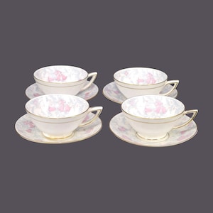 Op de afbeelding: Set van vier witte theekopjes en schoteltjes met een bloemenpatroon in roze en blauwe tinten. De kopjes en schoteltjes hebben een gouden rand.