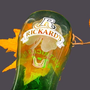 May include: A green glass bottle with a label that says "Rickard's" and a shamrock design. The bottle is partially filled with a yellow liquid and is sitting on a brown surface.