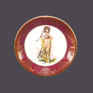 May include: A decorative plate with a red background and gold trim. The center of the plate features a painting of a young girl in a white dress and a pink hat. The girl is holding a pink ribbon in her hand.