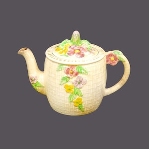 Könnte beinhalten: Eine cremefarbene Teekanne mit einem gewebten Muster und floralen Akzenten. Die Teekanne hat einen Deckel mit einem floralen Design und einen Henkel mit einem floralen Akzent.