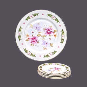 May include: A set of four white ceramic plates with a floral design featuring pink and purple roses, green leaves, and a gold trim.