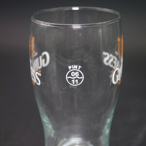 May include: A clear glass pint glass with the words "Guinness" printed on both sides. The glass has a white circle with the words "Pint 06 11" printed on it.