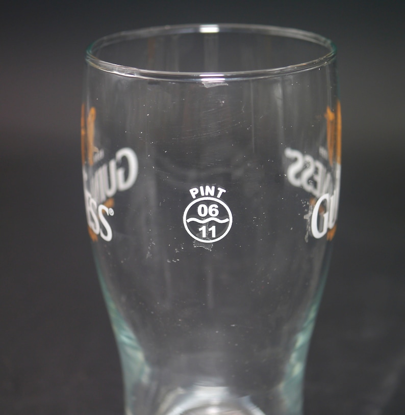 May include: A clear glass pint glass with the words "Guinness" printed on both sides. The glass has a white circle with the words "Pint 06 11" printed on it.