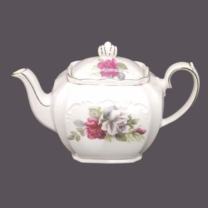May include: White porcelain teapot with a floral rose design in red, pink, and grey. The teapot has a gold trim, a curved spout, and a decorative lid with a small crown-like finial. The handle is also curved.