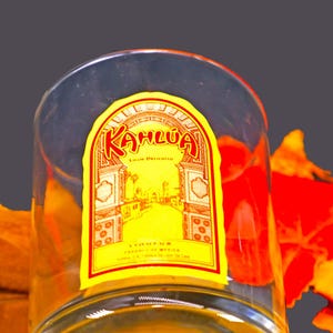 May include: A clear glass with a yellow and red Kahlúa label. The label features the brand name in red script, a yellow background, and an illustration of a building. The glass is partially filled and set against a grey background with orange and red leaves.