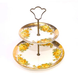 May include: Two-tiered serving stand with a gold rim and yellow rose pattern. The stand has a metal handle and is perfect for displaying desserts or snacks.