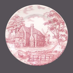 May include: A white plate featuring a red transferware design of a rural scene with a house, windmill, trees, and figures. The plate has a decorative border and is suitable for serving food. The design is reminiscent of traditional tableware.