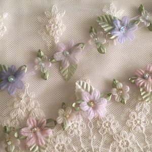 May include: A delicate floral trim with pastel pink, purple, and white flowers and green leaves. The trim is made of fabric and has a satin ribbon woven through it.